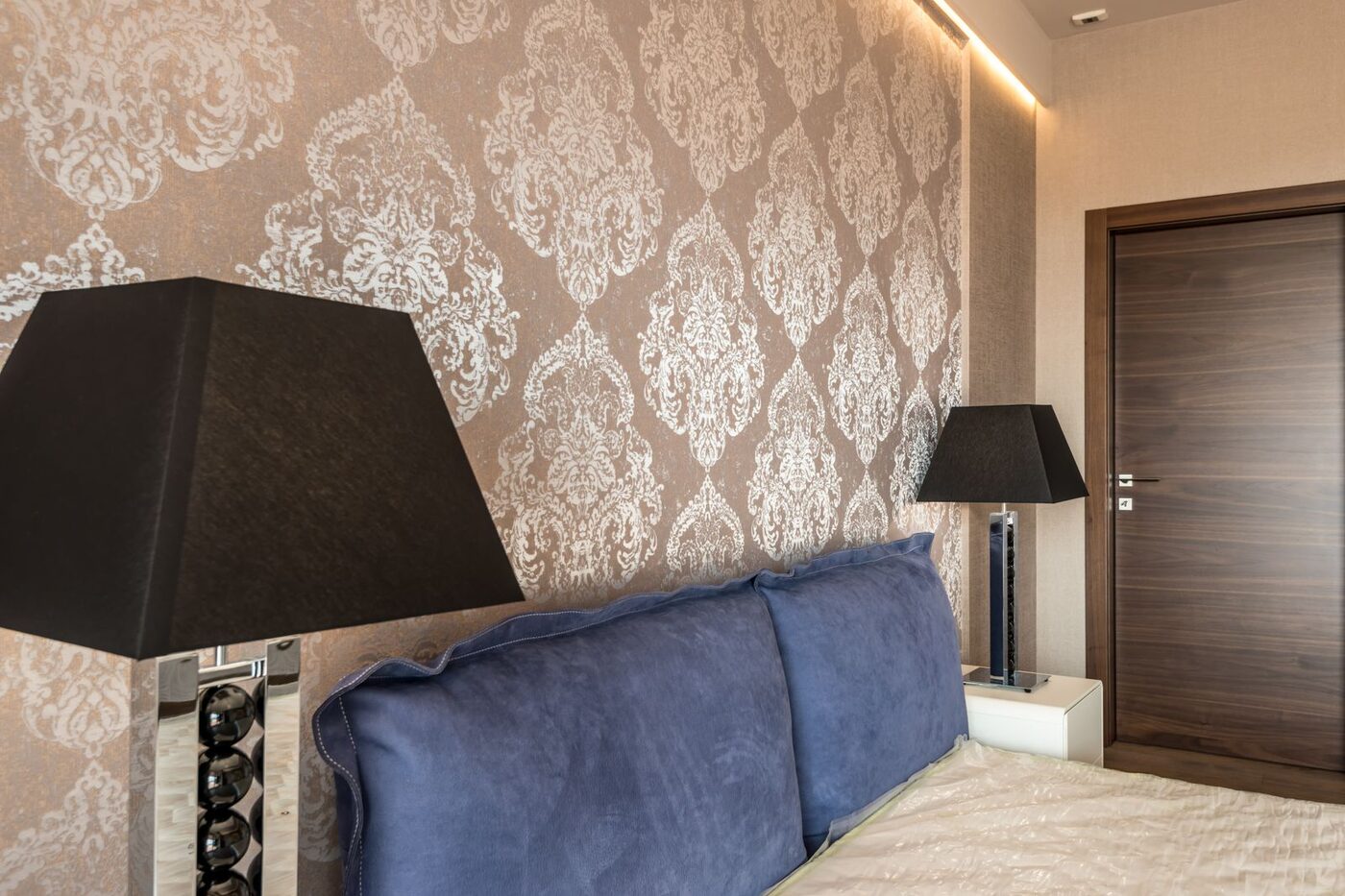 A bedroom with a beige damask wallpaper feature wall, dark lamps and a paneled wood door