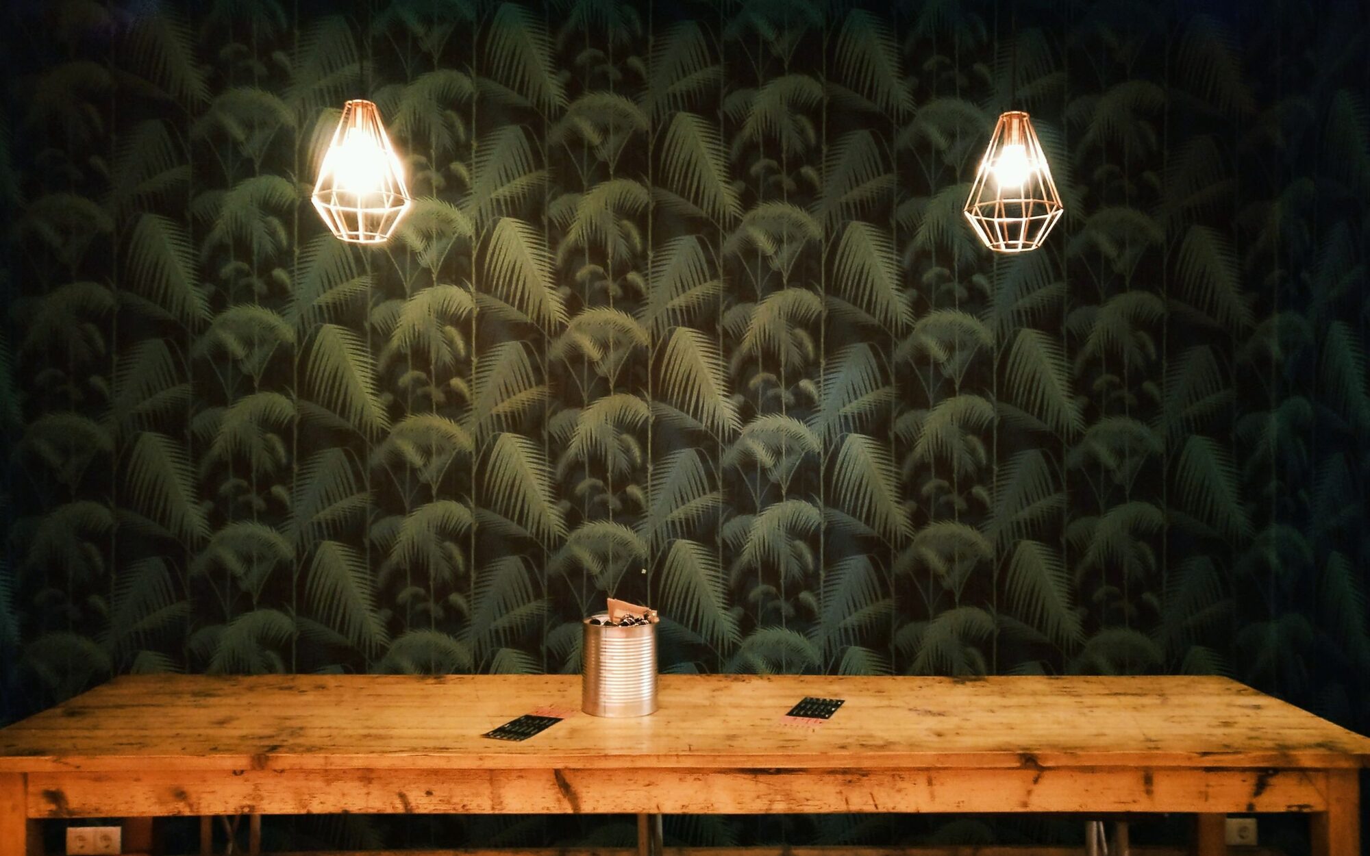 A dining space with a dark botanical mural wallpaper behind a rustic wood table lit by two copper cage pendants